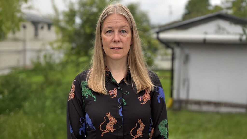 Kvinna med blont hår, svar blus. står utomhus